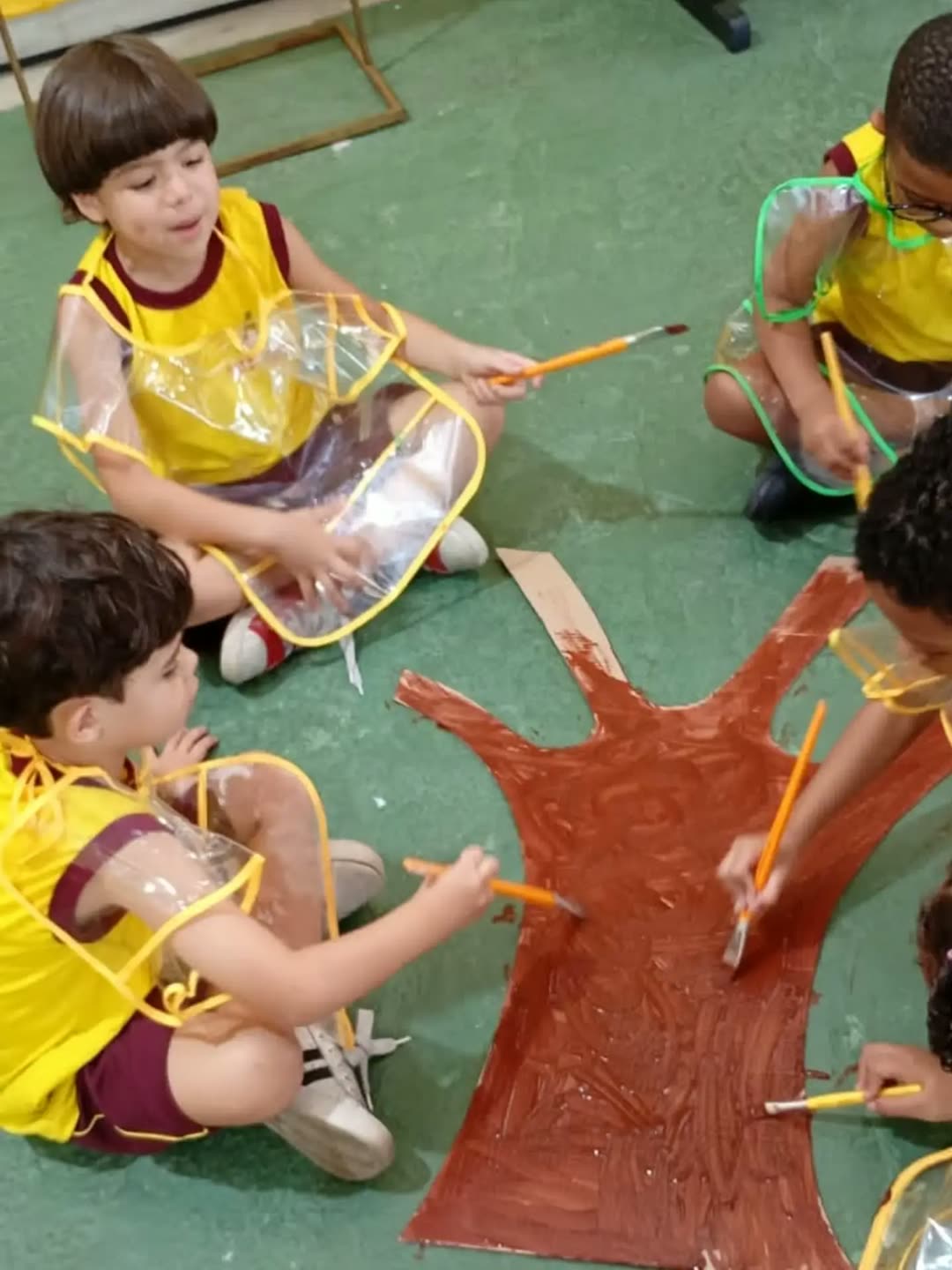 Crianças de uniforme amarelo e vinho pintando de marrom o tronco de uma árvore de papelão no pátio da escola