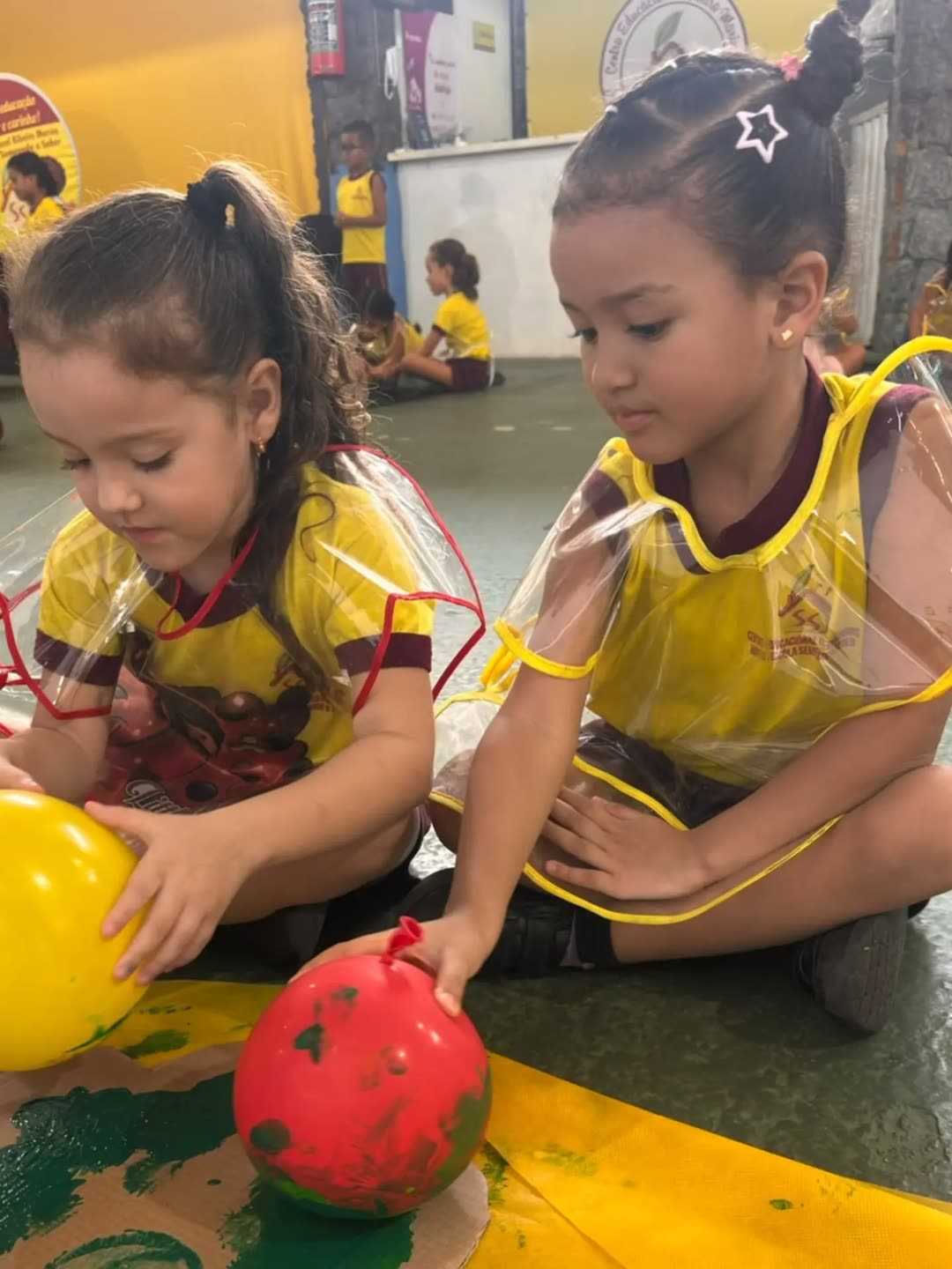 Duas meninas de uniforme carimbando a árvore com bolinhas sujas de tinta verde
