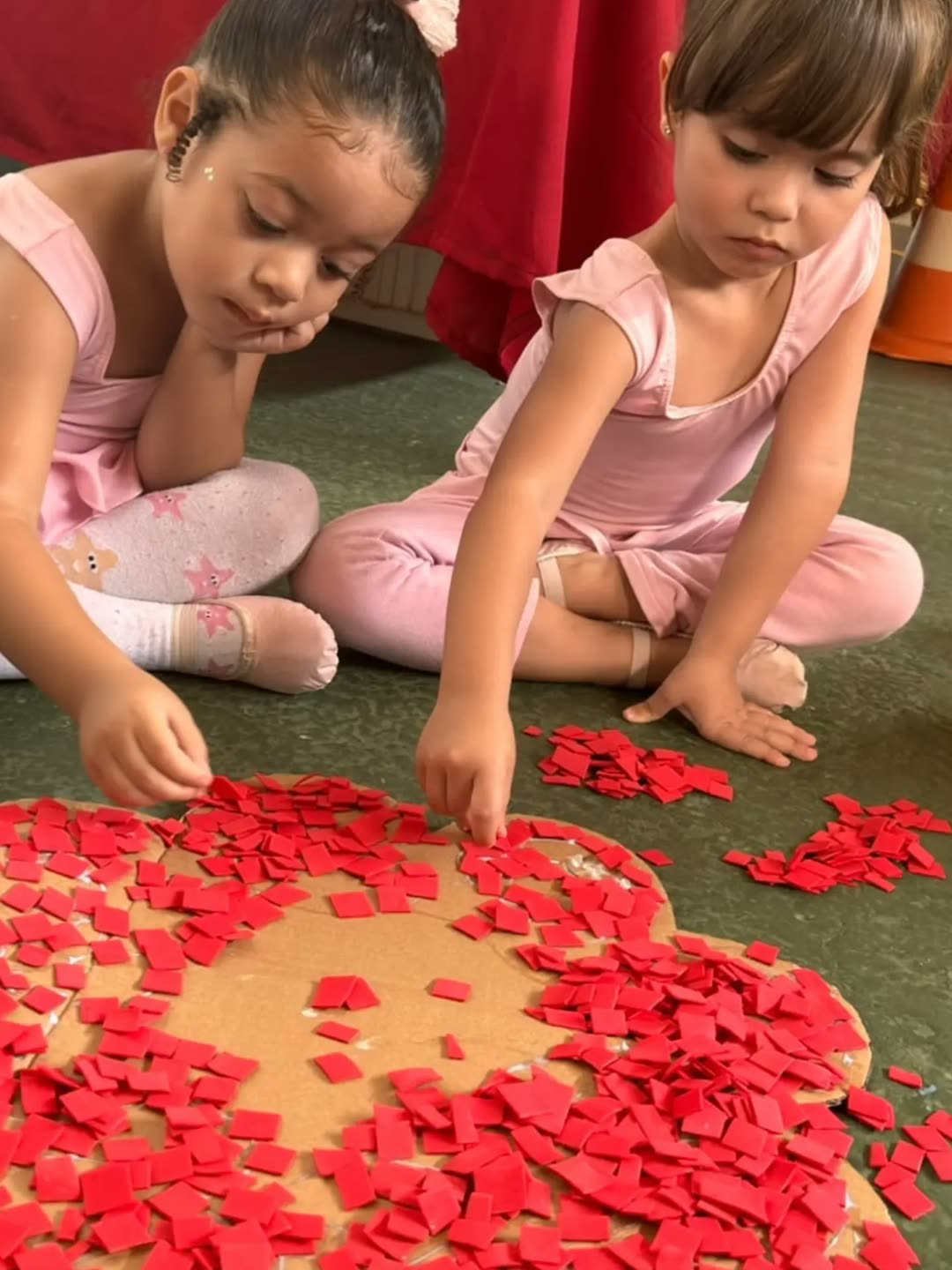 Duas meninas de roupa de ballet colando pedacinhos de papel vermelho numa flor de papelão