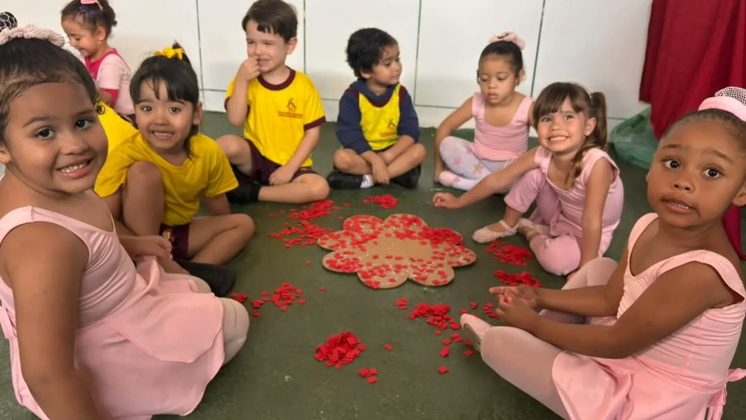 Turma sentada em roda no chão da escola montando uma flor de papel vermelho, várias crianças olhando e sorrindo para a câmera